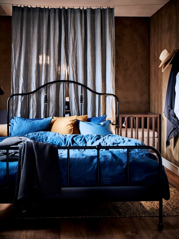 Un dormitor compartimentat subtil cu două perdele cu un cadru de pat SAGSTUA din metal negru în mijlocul camerei și un pătuț.