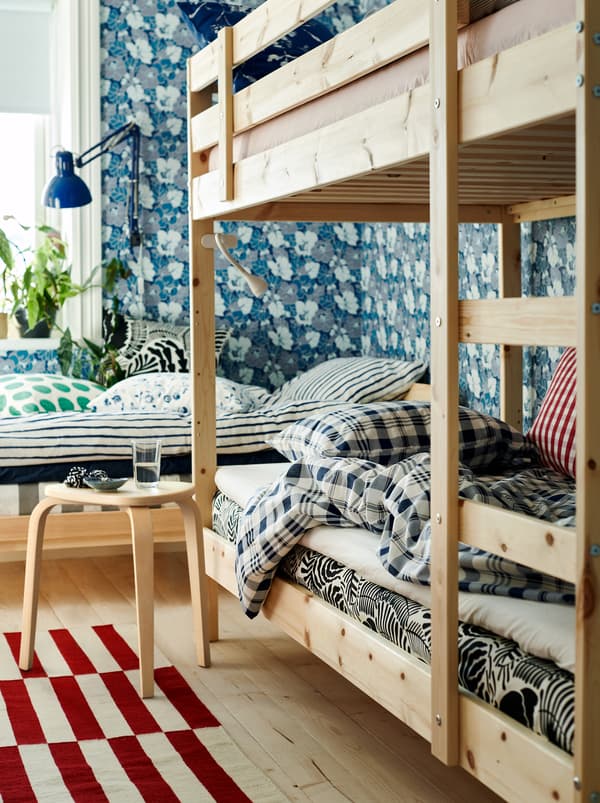 Un dormitor cu pat și pat supraetajat în culori neutre, tapet cu model floral și textile în culori și modele diferite.