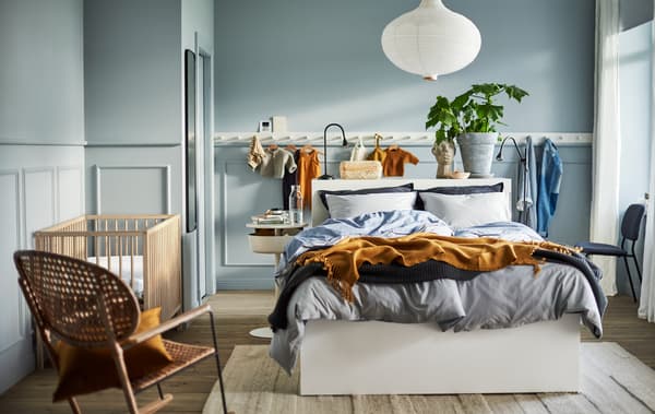 Un dormitor în albastru deschis, cu un pat alb, un corp de iluminat alb, un panou alb cu cârlige, un balansoar din ratan și un pătuț SNIGLAR.