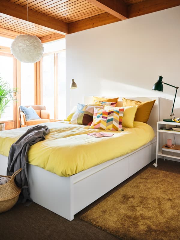 Un dormitor însorit și luminos cu un pat MALM alb, acoperit cu o cuvertură galben intens și perne HANNELISE în mijloc.