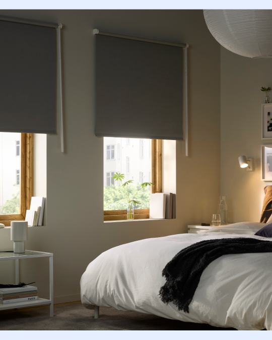 Un dormitorio con dos ventanas que tienen cortinas rollar blackout a medio bajar impidiendo que la luz del sol llegue al cabecero de la cama.