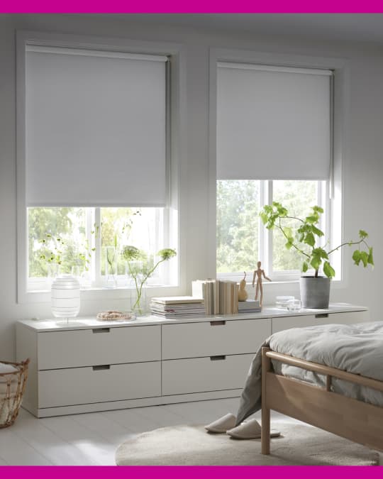 Un dormitorio con una cama de 2 plazas y en la ventana hay dos cómodas grandes. Un par de cortinas roller black out a medio cerrar cubren la luz.