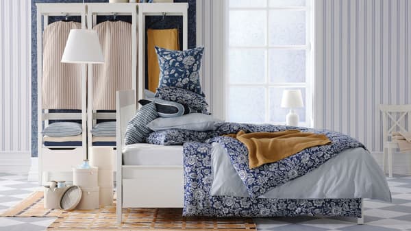 Un dormitorio en el que aparece una cama de estructura blanca en primer plano con funda nórdica y cojines a juego de estampado de flores blancas y azul. Alfombra de estampado geométrico amarillo y banco, lámpara y cajas blancas y al fondo un armario con ropa, una ventana y pared de papel pintado de rayas verticales.