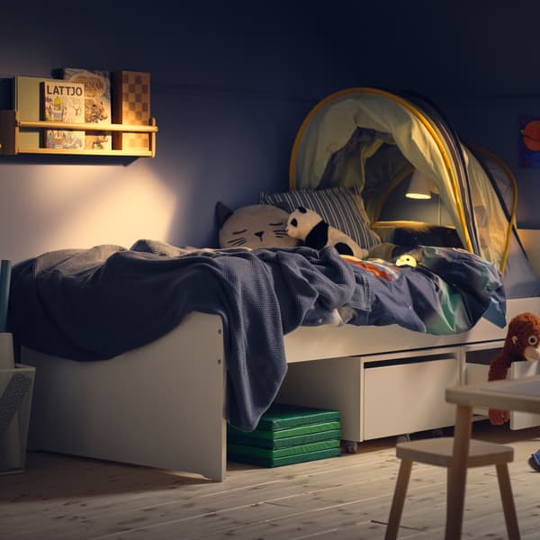 Un dormitorio infantil azul, iluminado de xeito tenue, cunha cama SLÄKT branca con caixas de almacenaxe con rodas debaixo e un dosel ELDFLUGA enriba.