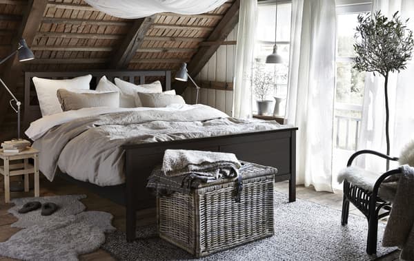 Un dormitorio rústico cunha estrutura de cama HEMNES negra-marrón, unha cesta de vimbio, pel de ovella no chan e teitos de madeira en mansarda.