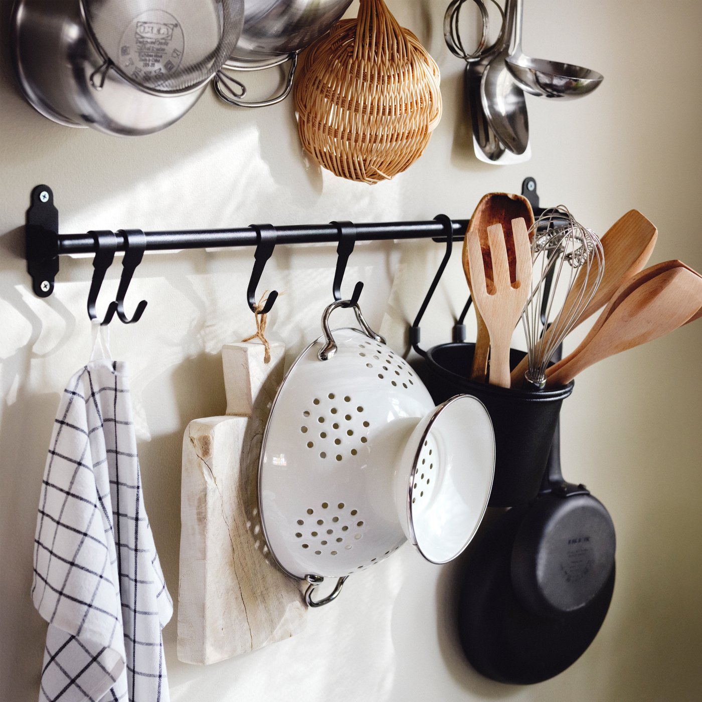 Un drap de cuina, una fusta de tallar, un escorredor blanc i utensilis en un recipient HULTARP negre penjat en una guia HULTARP negra.