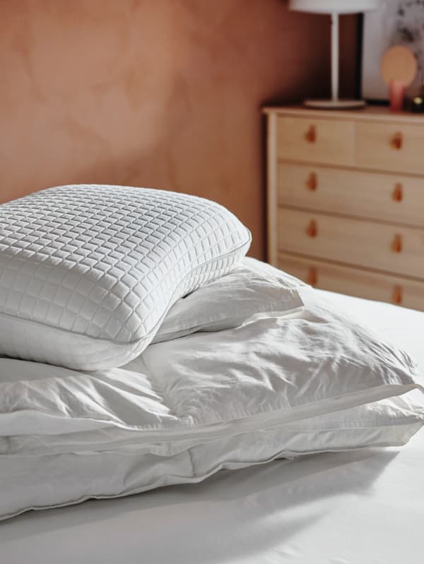 Un edredón FJÄLLARNIKA e unha almofada ergonómica KLUBBSPORRE amontoadas sobre a cama nun dormitorio iluminado polo sol.