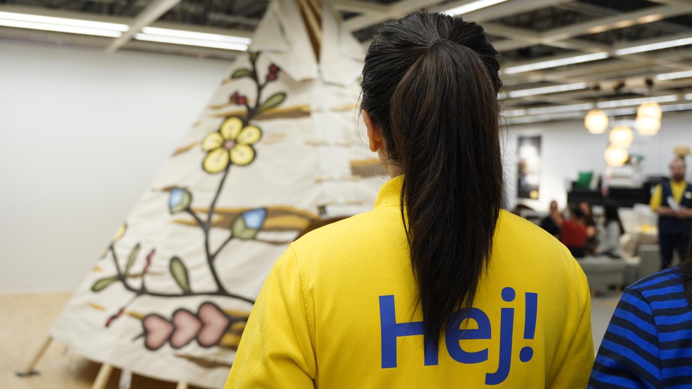 Un employé d'IKEA, vêtu d'une veste jaune portant l'inscription « HEJ ! » en bleu dans le dos, se tient debout en regardant un tipi dans un magasin IKEA.