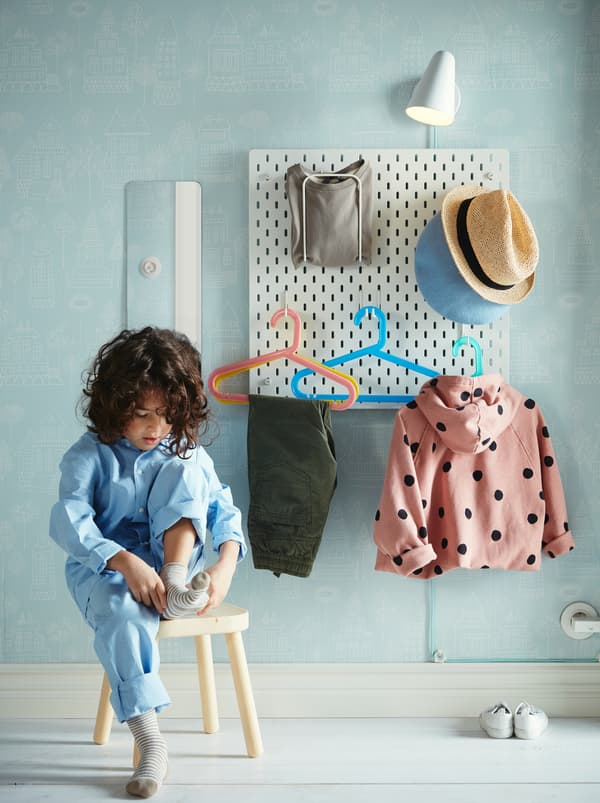 Un enfant assis sur un tabouret qui enfile des chaussettes devant un panneau perforé SKÅDIS où sont suspendus divers articles vestimentaires.