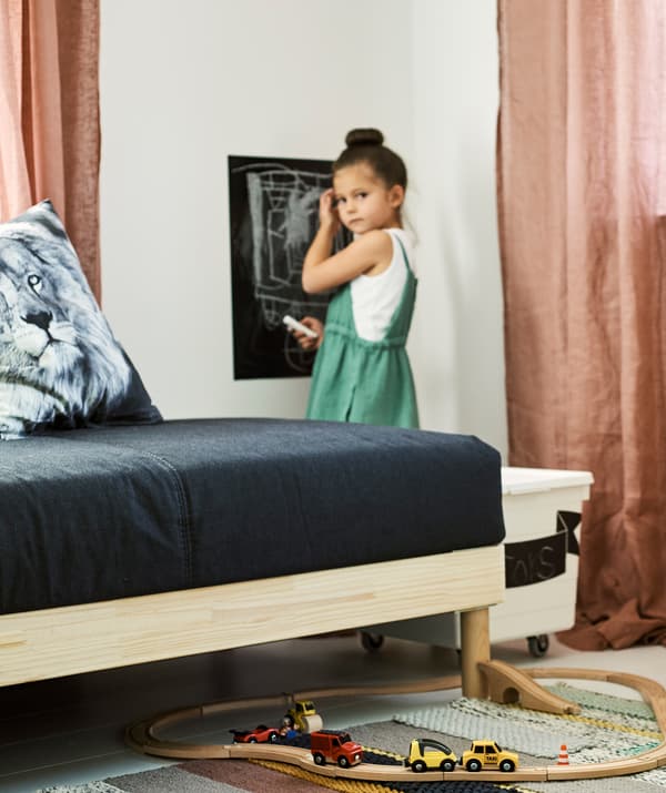 Un enfant dessine sur un autocollant mural en peinture pour tableau noir dans une pièce avec une boîte à jouets, des rideaux roses, un canapé gris et un petit train en jouet sur un tapis à rayures.