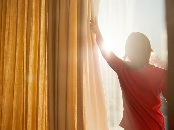 Un enfant en t-shirt rouge ouvre les rideaux jaunes et blancs d’une fenêtre, laissant passer les rayons du soleil.