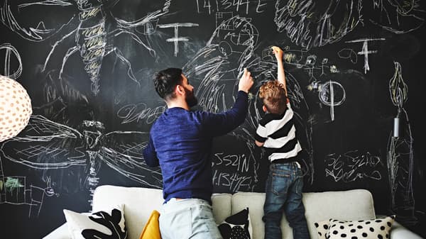 Un enfant et un adulte, l’un debout, l’autre agenouillé, sur un canapé, en train de dessiner sur un mur transformé en tableau noir. Des coussins de différentes couleurs sont dispersés sur le canapé.