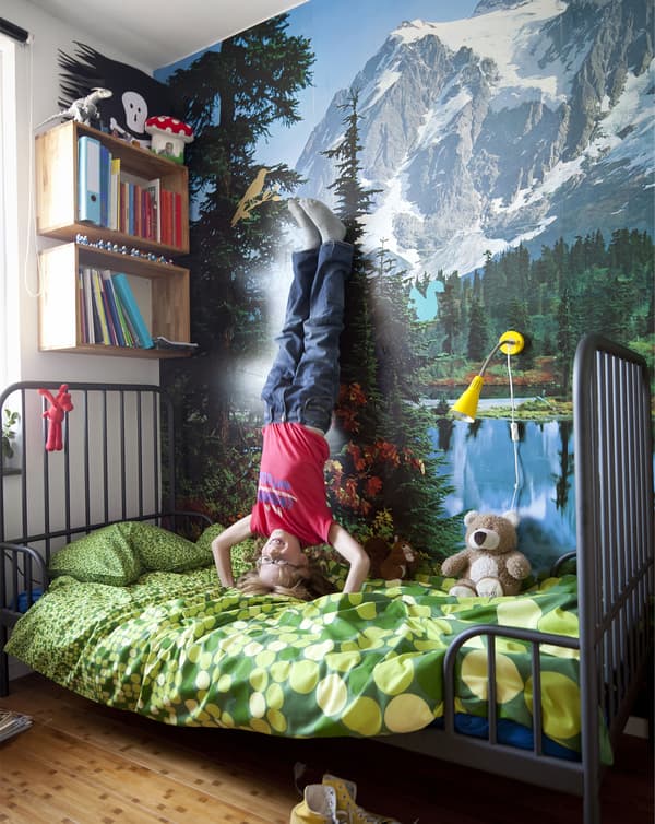 Un enfant fait le poirier sur un lit et pose ses pieds contre un mur décoré d’un paysage de montagne.