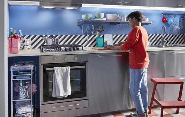 Un enfant prépare une collation dans une cuisine en acier inoxydable avec des étagères et un marchepied.