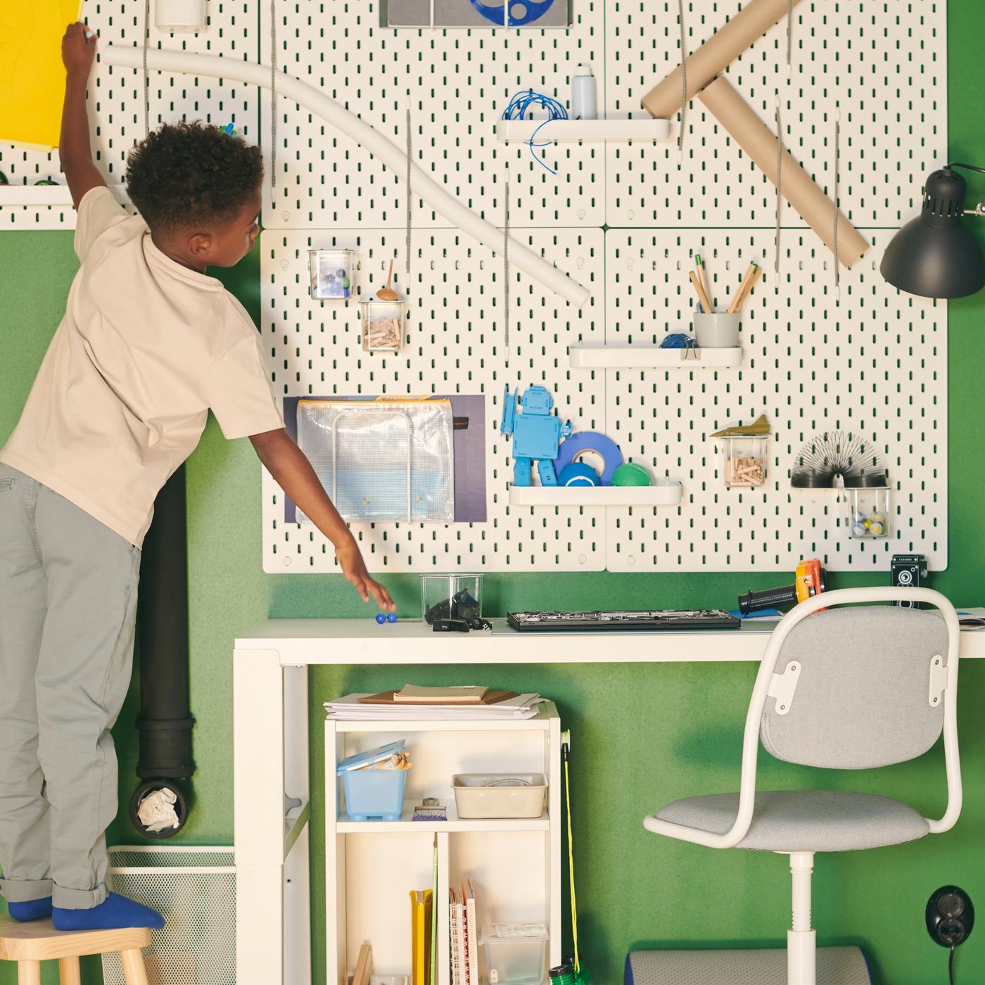 Un enfant tend les mains vers le mur avec des panneaux perforés blancs avec divers articles, au-dessus d'un bureau blanc avec une chaise de bureau pour enfants.
