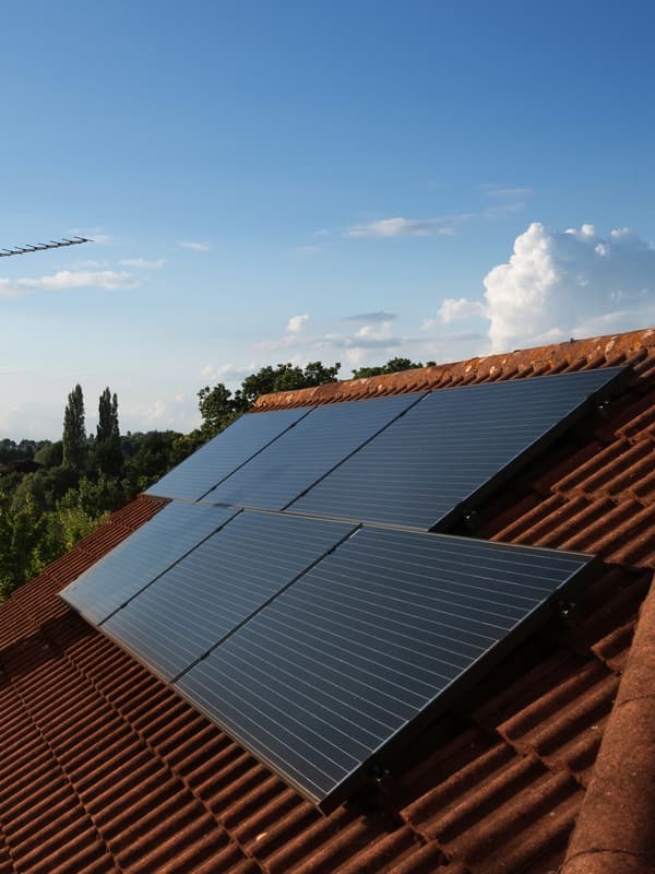 Un ensemble de six panneaux solaires sur un toit en pente, recouvert de tuiles en terre cuite rouge. À l’arrière-plan, des arbres et un ciel bleu.