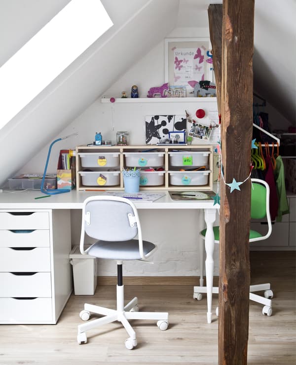 Un escritorio infantil con caixas de almacenaxe e dúas cadeiras de oficina nun dormitorio branco.