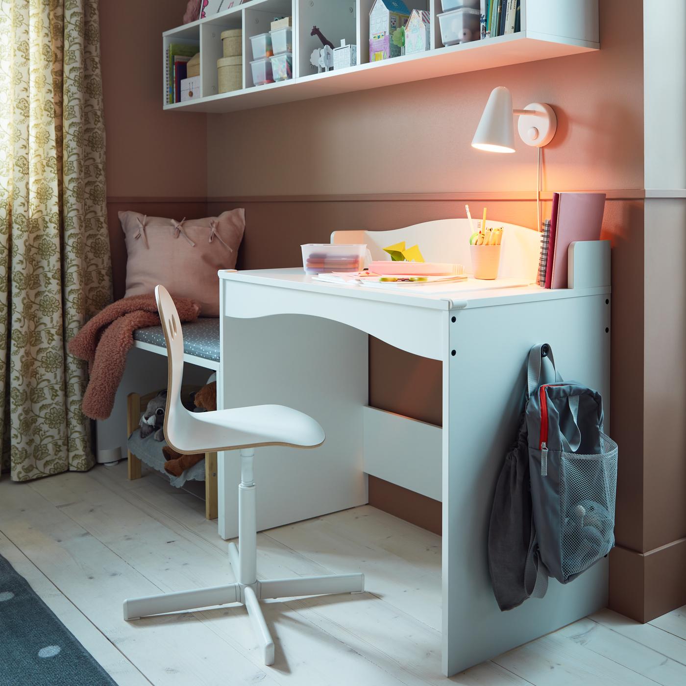 Un escritorio SMÅGÖRA blanco con dos bolsas en un gancho, una silla de escritorio infantil blanca, una lámpara de pared LED blanca y dos estanterías blancas.