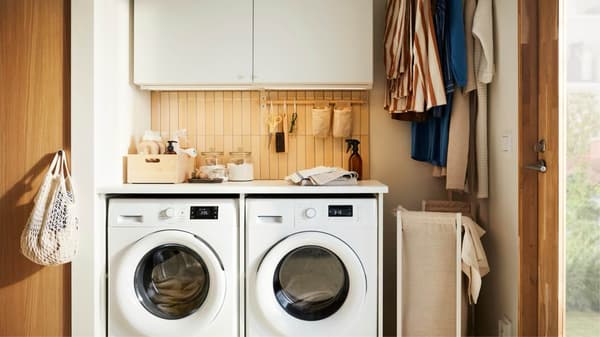Un espace buanderie compact ENHET présente deux machines à laver sous un plan de travail, un panneau mural en bois, des rangements et des vêtements suspendus sur le côté.