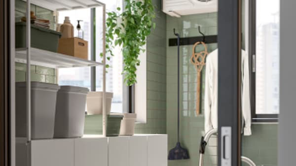 Un espace de rangement lumineux et moderne présente des étagères ouvertes JOSTEIN avec des boîtes, une plante suspendue et des accessoires de nettoyage près d’une fenêtre.