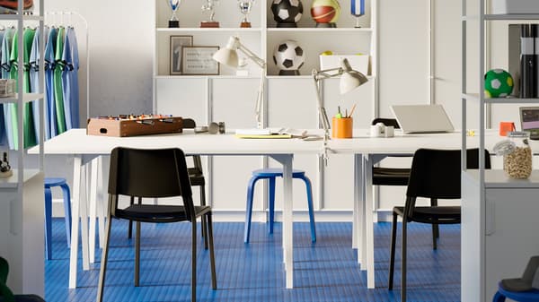 Un espace de réunion ouvert avec des tables TROTTEN blanches, des chaises noires et une bibliothèque BILLY dans un bureau au sol bleu et aux murs blancs