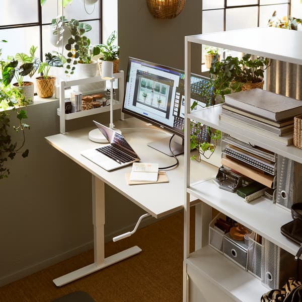 Un espace de travail centré sur un bureau assis-debout TROTTEN niché entre un rangement à portes coulissantes TROTTEN blanc et une fenêtre.