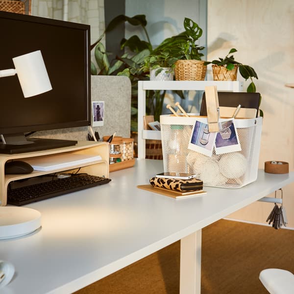 Un espace de travail faiblement ensoleillé avec un bureau assis-debout TROTTEN, un support pour ordinateur portable VATTENKAR et un panier RISATORP contenant des accessoires.