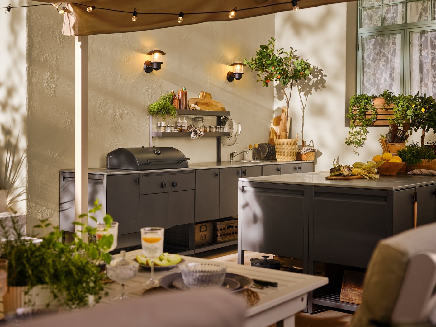 Un espace extérieur contient une table BONDHOLMEN blanc/beige et plusieurs modules BÅTSKÄR gris foncé, dont un barbecue au charbon.