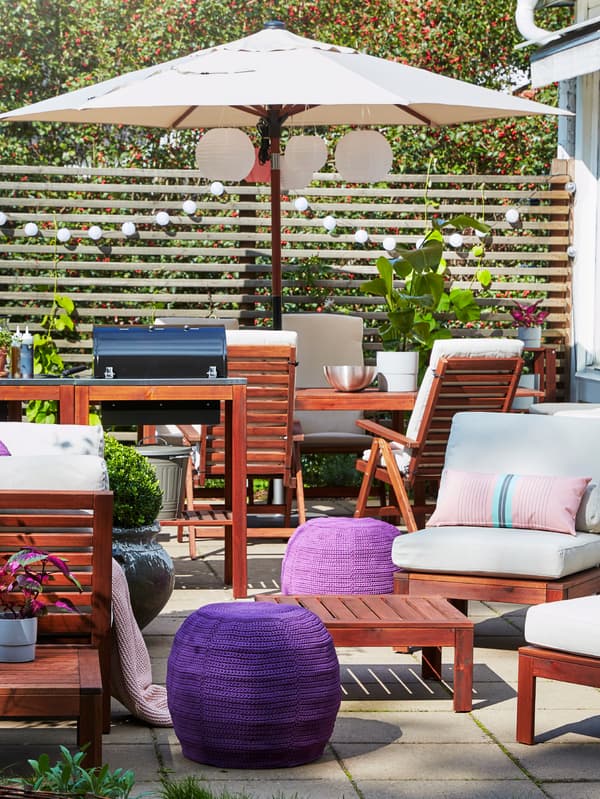 Un espace extérieur, meublé avec des tables et des chaises d’extérieur, un parasol, et agrémenté de coussins, de poufs et de luminaires.