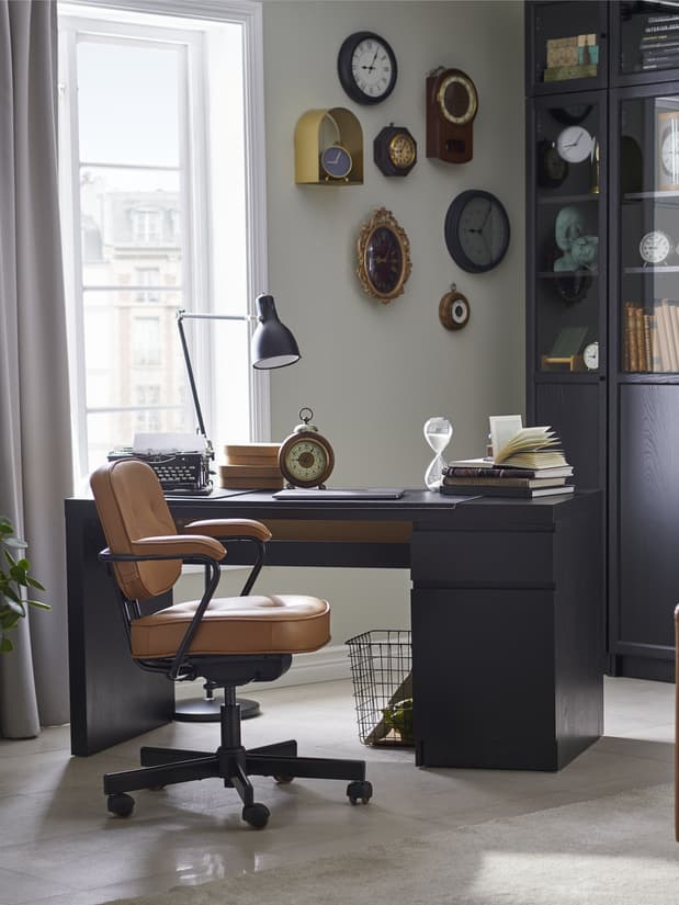 Un espacio de trabajo con un escritorio MALM negro una silla de escritorio elegante café. En el ondo se ven diversos relojes en la pared y una vitrina con decoración