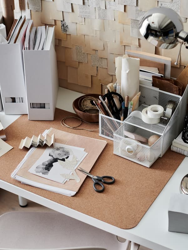 Un espacio de trabajo luminoso y algo desordenado con un organizador de escritorio DRÖNJÖNS, un protector de escritorio SUSIG de corcho y una lámpara FORSÅ niquelada.