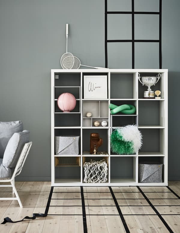 Un estante KALLAX de IKEA branco garda a decoración e caixas cunha parede gris/verde de fondo.