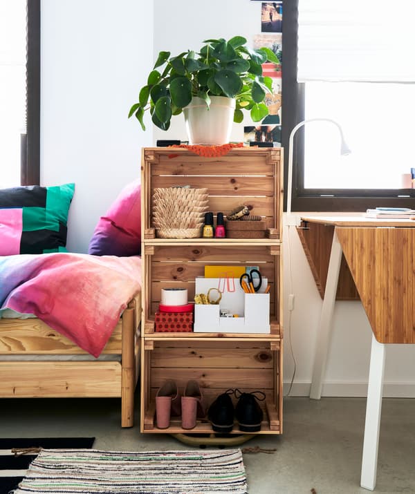 Un estante lixeiramente cheo con tres caixas de madeira colocadas ao carón e encasteladas entre unha cama e unha pequena mesa de comedor.