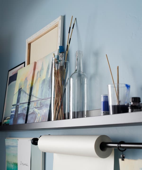 Un estante para cuadros utilizado para guardar herramientas de arte con una barra de cortina debajo en la que se ha fijado un rollo de papel para pintar.