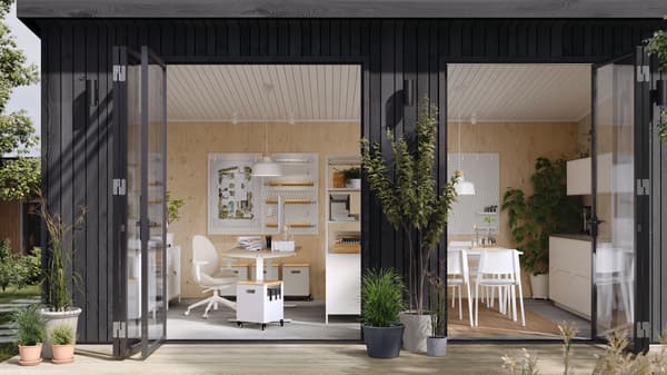 Un estudio en el jardín con puertas abiertas; en su interior hay una zona de trabajo con un escritorio y una silla, una estantería abierta como separador de espacios y una zona de cocina comunitaria.