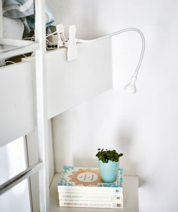 Un faretto bianco da parete con morsetto illumina una pila di libri su una mensola - IKEA