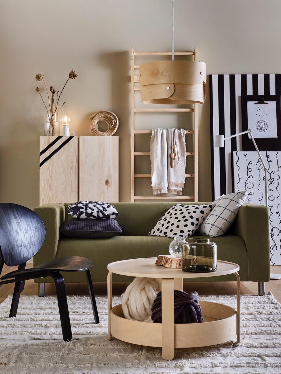 Un fauteuil noir et une table basse en bouleau sur un tapis naturel, à côté d’un canapé jaune-vert avec des meubles IVAR en pin derrière lui.