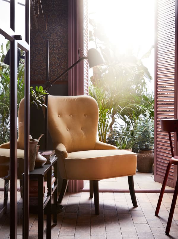 Un fauteuil REMSTA jaune installé devant une porte ouverte donnant sur une terrasse décorée de plantes. Un lampadaire noir et une petite table contenant des livres complètent l’ensemble.