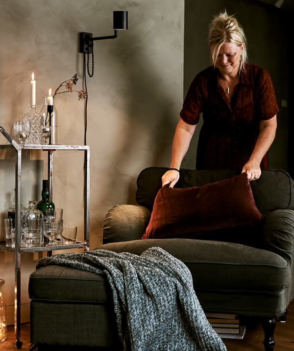 Un fauteuil vert à côté d’un chariot à boisson avec des verres, une femme tapote un coussin de velours rouge.