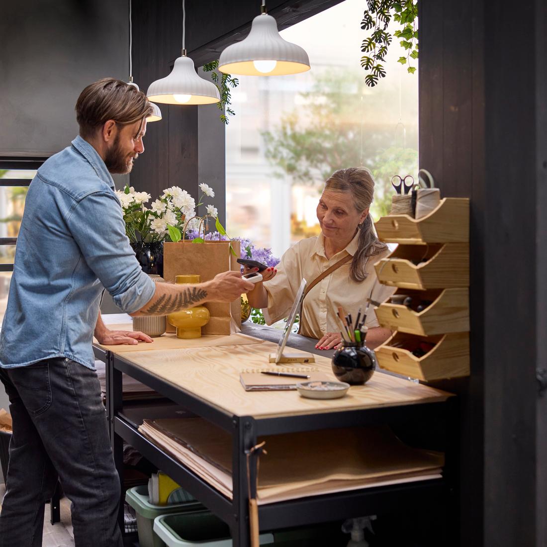 Un fleuriste aide un client à faire ses achats, avec en arrière-plan un établi BROR en contreplaqué noir et pin, entouré de plantes en pot et de fleurs.