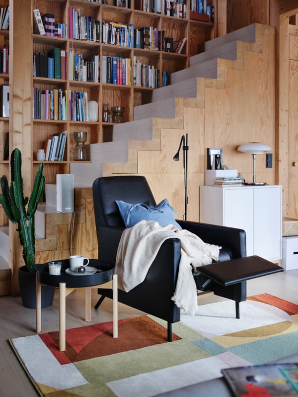 Un fotoliu rabatabil GISTAD negru acoperit cu pături se află într-o cameră cu pereți de lemn, cu sprijinul pentru picioare în poziție ridicată.