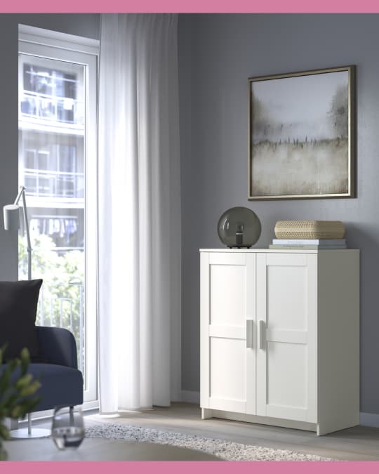 Un gabinete IKEA de una puerta apoyado en la pared al lado de la ventana de una sala.