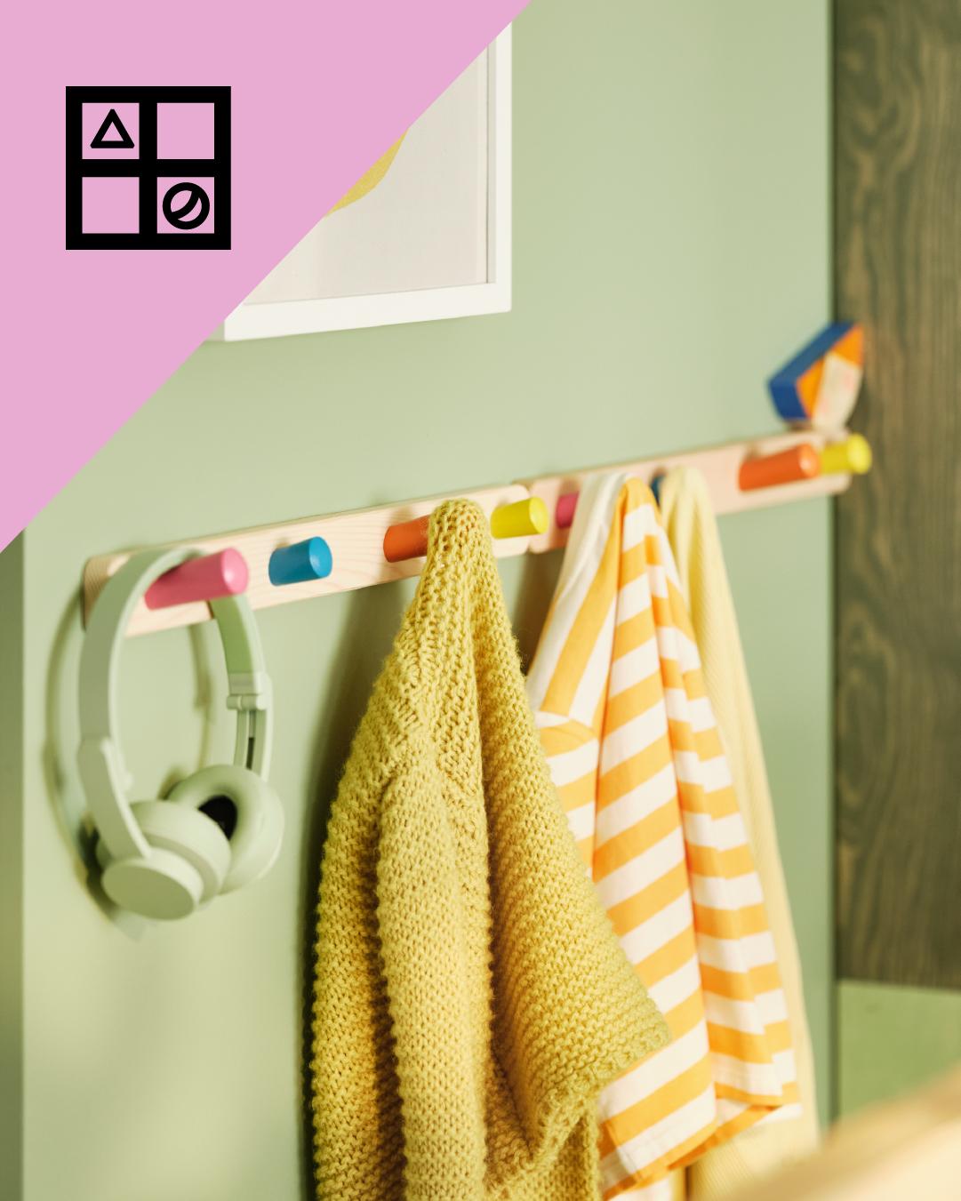 Un gancho de madera para colgar ropa de niños con perchas de colores del que cuelgan unos audífonosy dos chaquetas de niños. En la parte superior de la imágen triángulo rosa con figuras geométricas.