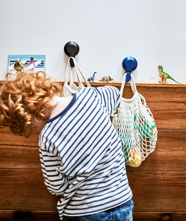 Un garçon cherche un jouet dans un filet suspendu à un crochet à côté d'un deuxième filet, sur fond de mur blanc garni de panneaux bois.