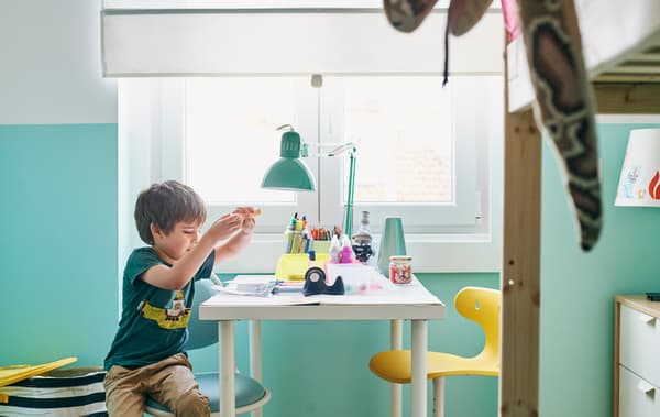 Un garçon est installé à un bureau, sur une chaise blanche et turquoise. Sur le bureau, des feutres et une lampe de bureau verte, et de l’autre côté, une chaise jaune.