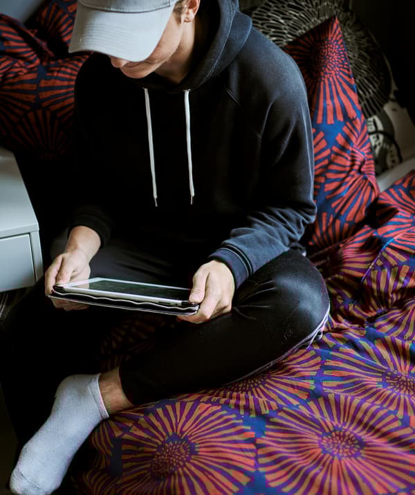 Un garçon portant une casquette de baseball joue sur une tablette, assis sur un lit avec une couette aux imprimés graphiques rouges et bleus.