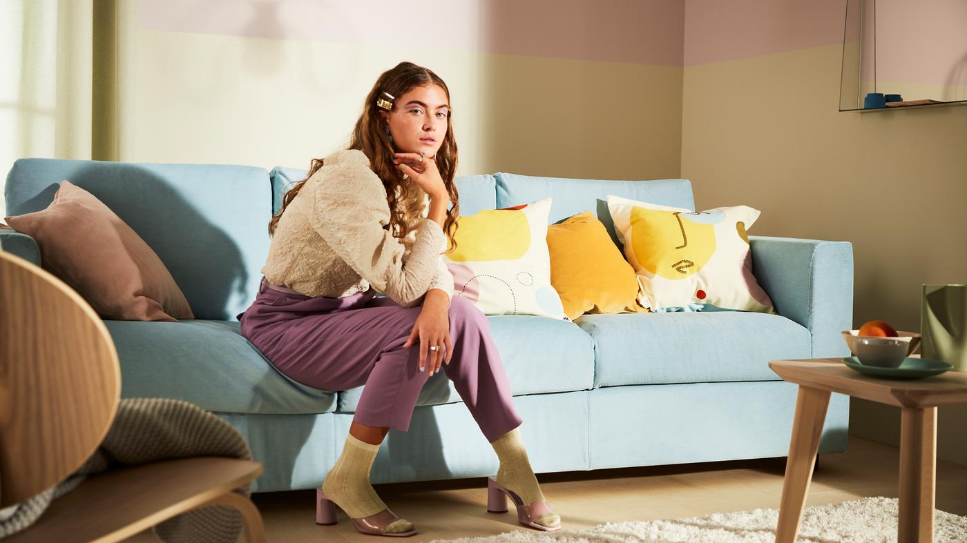 Un grand canapé bleu clair avec différents coussins et une jeune femme assise sur le canapé dans un salon moderne aux couleurs pastels.