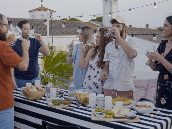 Un groupe de jeunes, debout devant une table de buffet pleine de nourriture sur une terrasse extérieure, mangent, boivent et discutent dans la joie et la bonne humeur.