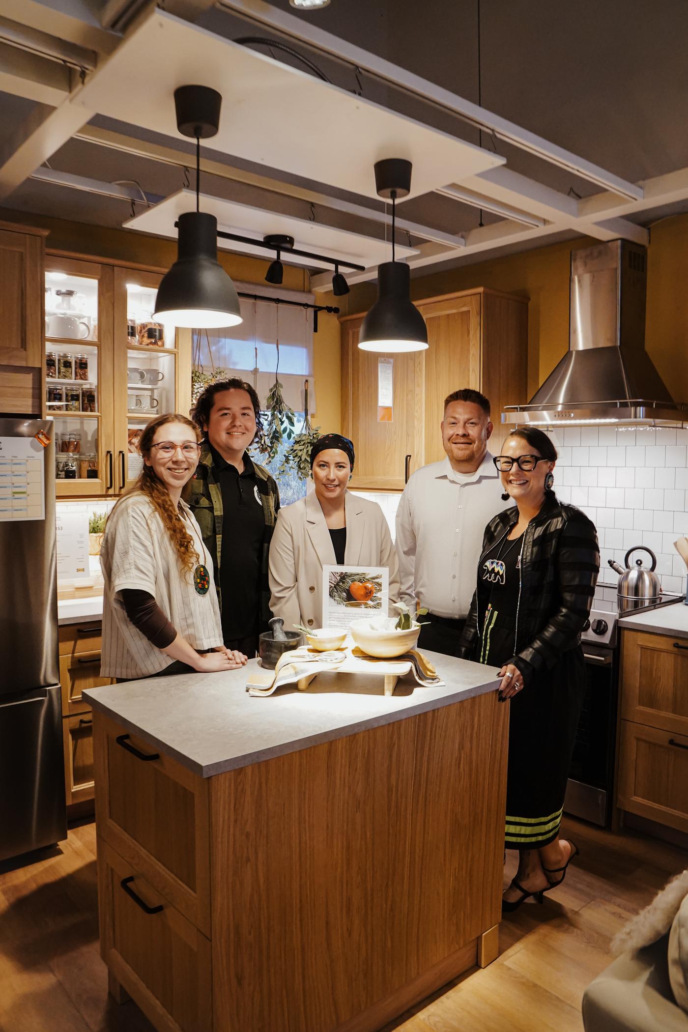 Un groupe de personnes rassemblé autour d’un îlot de cuisine regardent la caméra en souriant.