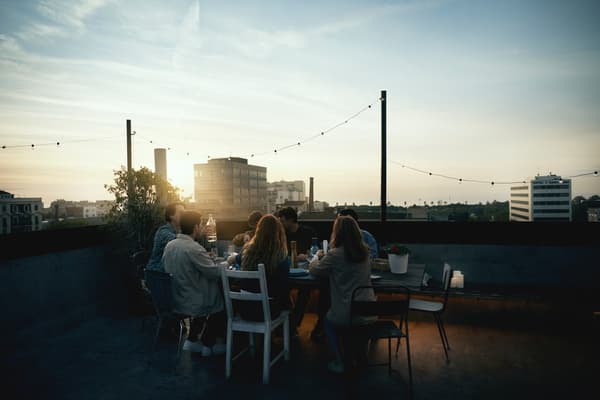 Un grupo de personas disfrutando en la azotea de un edificio, sentados todos al rededor de una mesa y con sillas de diferentes colores y estilos. Todo esto mientras el sol se pone.